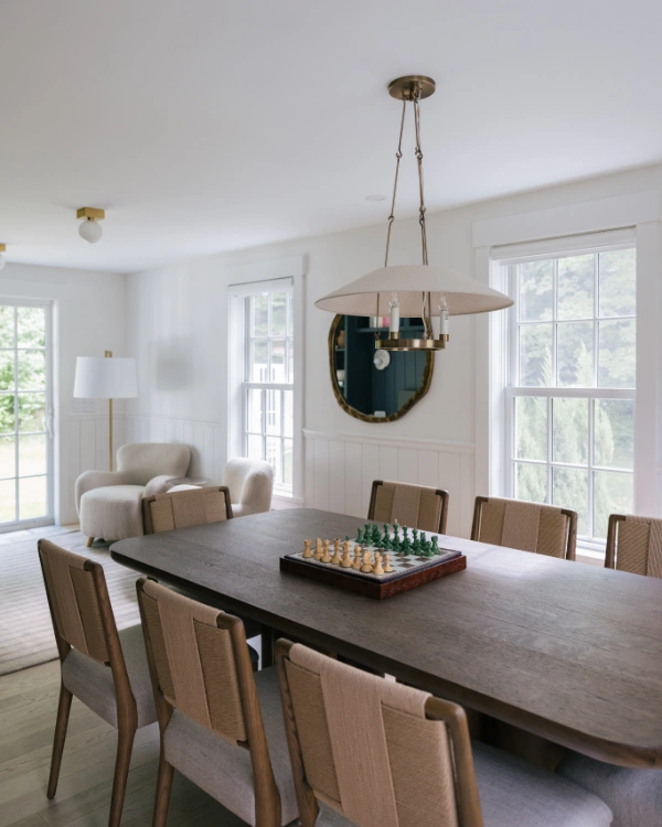 Dining table setup with upholstered chairs, oversized brass pendant light, and chess board centerpiece beneath tall windows